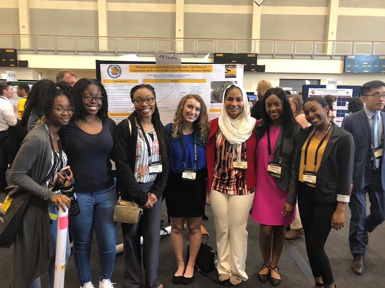 Seven students at a research poster titled Wild and Commercial Mushroom Cultivation at a conference