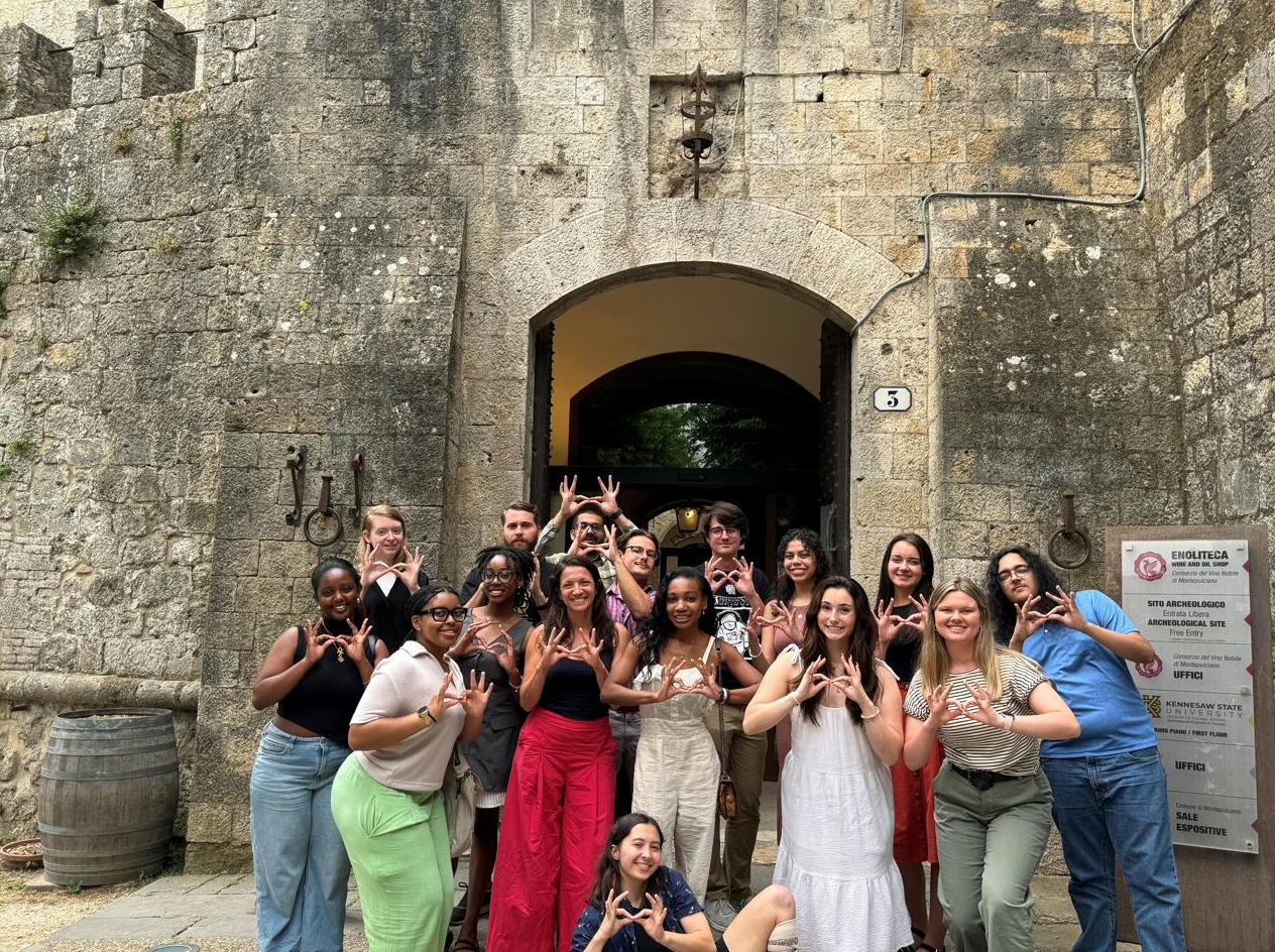 A group of students posing with hand signs outside a stone archway in Montepulciano, Italy